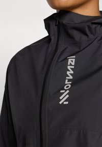 Schwarze wasserdichte Jacke mit hohem Kragen, Reißverschluss und reflektierendem Logo auf der Vorderseite. Glatte, leichte Stofftextur.