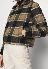 Chaqueta de forro polar con capucha, con un patrón de cuadros en blanco y negro y cierre frontal de cremallera. Tejido grueso y texturizado con puños elásticos.