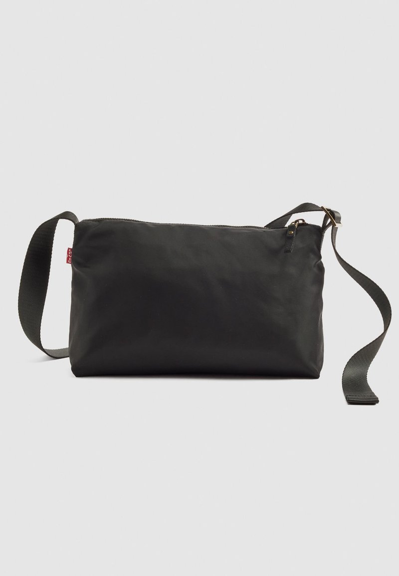 Bolso cruzado rectangular negro con correa ajustable, cierre de cremallera y pequeña etiqueta de marca roja en el lateral sobre fondo liso.