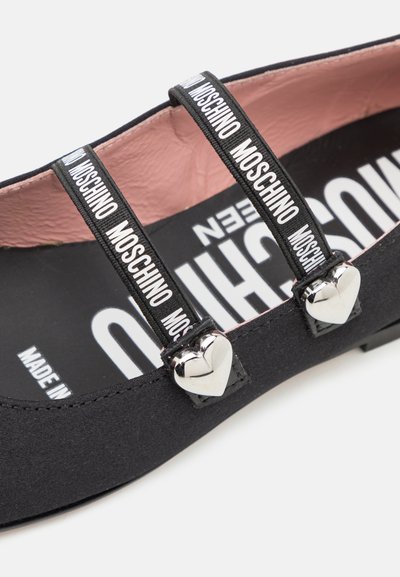 Zapato plano negro hecho de tela suave, con dos correas con la marca "MOSCHINO", acentos de corazón plateado y un forro interior rosa.