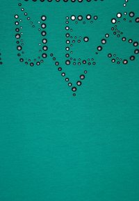 Tela verde azulado con textura suave, decorada con adornos plateados en forma de puntos, formando letras hechas de lentejuelas redondas.