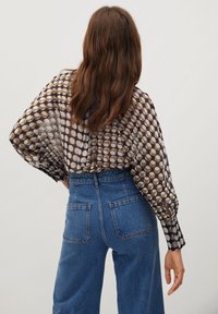 Blusa marrón transparente con patrones circulares, mangas holgadas y espalda redondeada. Combinada con unos jeans de mezclilla azul de tiro alto.