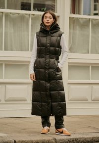 Vesta puf din material negru cu glugă, având secțiuni matlasate vertical și orizontal, talie strânsă și buzunare laterale, purtată cu pantaloni gri și adidași portocalii.