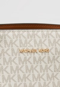 Flødefarvet tekstureret overflade med gentaget lysebrunt "MK" mønster og guld "MICHAEL KORS" logo, accentueret af en brun læderkant med syninger.