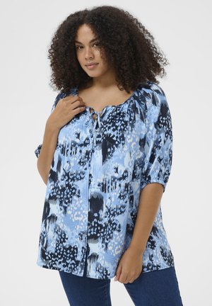 Femme aux cheveux bouclés portant un chemisier bleu ample à motifs avec un col noué et des manches mi-longues, associée à un jean bleu foncé.