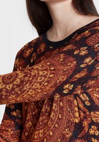 Camisa de manga larga con un fondo oscuro, que presenta intrincados patrones de paisley en naranja y dorado. Textura suave con un escote redondo.