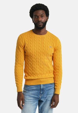 Geel gebreid sweater met een kabelpatroon, ronde halslijn en geribde manchetten en zoom. Bevat een klein geborduurd logo op de borst.