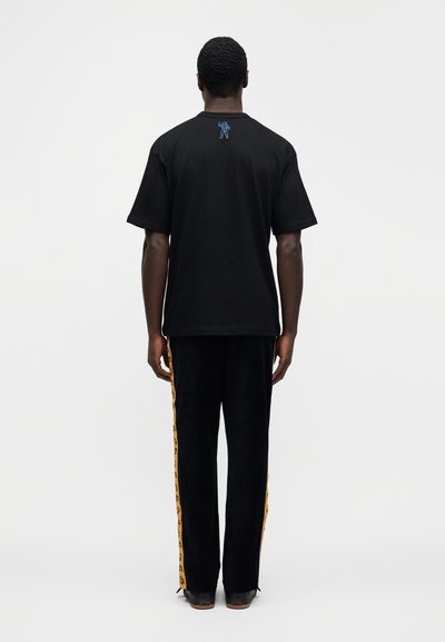 Μαύρο βαμβακερό T-shirt με κοντά μα νίκια, με μπλε κεντημένη λεπτομέρεια στην πλάτη. Συνδυάζεται με μαύρο παντελόνι που έχει κίτρινες ρίγες στο πλάι.