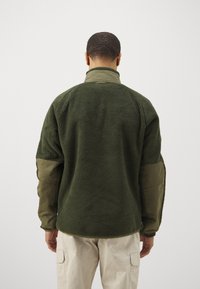 Jachetă de fleece verde cu guler înalt și mâneci de un verde măsliniu contrastant. Prezintă o croială lejeră și un material texturat.