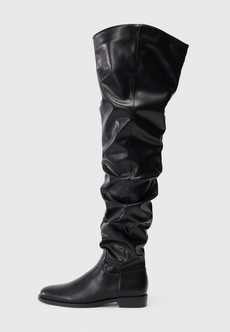 Botas negras por encima de la rodilla hechas de cuero suave. Presentan un diseño holgado con suelas planas, puntas redondeadas y un hardware mínimo.