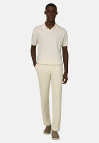 Homme portant un polo en maille à manches courtes crème, un pantalon crème assorti et des chaussures marron sans lacets, debout devant un fond clair uni.