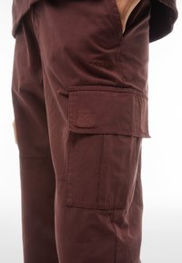Pantaloni cargo bordeaux con tasca laterale, realizzati in tessuto leggero. Presentano un logo cucito sottile e un design minimalista senza hardware visibile.