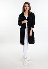 Cardigan negru tricotat cu un model în cablu, cu deschidere în față și mâneci lungi, purtat peste un top alb și pantaloni albi, asortat cu teniși albi.