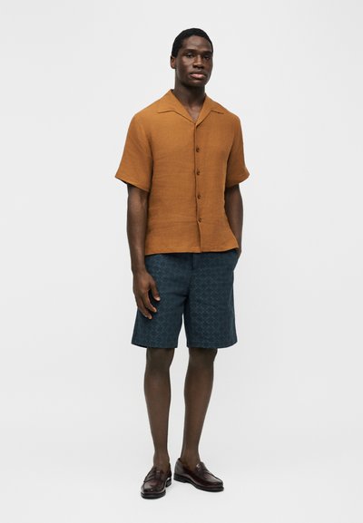 Άνδρας με κοντομάνικο πουκάμισο σε σκουριά απόχρωση, μπλε σκούρες σορτς με σχέδια μέχρι το γόνατο και σκούρα καφέ δερμάτινα μοκασίνια, στέκεται μπροστά σε λευκό φόντο.