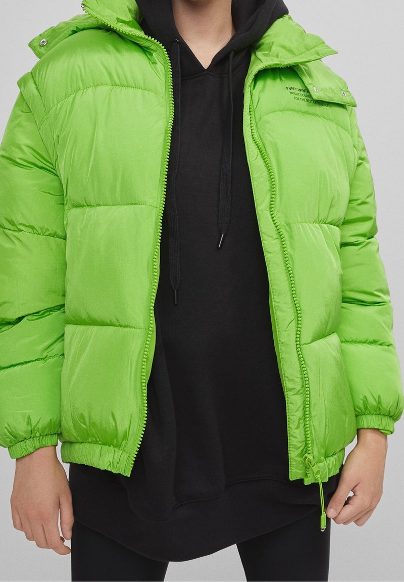 Doudoune verte avec un matériau léger et brillant, fermeture éclair et capuche. Portée sur un sweat à capuche noir, avec des poignets et un ourlet élastiques.