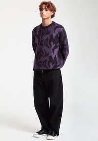 Suéter de patrones morados y negros con cuello redondo, combinado con pantalones de pierna ancha negros y zapatillas negras con suelas blancas.