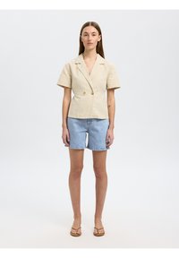 Blazer beige à rayures, à manches courtes et à double boutons, associé à un short en denim bleu clair. Le modèle se tient debout, portant des sandales marron.