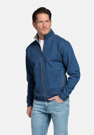 Man in blauw jack met rits en hoge kraag en lichtblauwe spijkerbroek, staand met één hand in de zak, kijkt iets naar rechts.