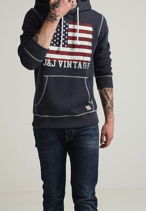 Osoba w ciemnoniebieskiej bluzie z kapturem z flagą amerykańską i napisem „J&J VINTAGE”, ciemnych jeansach i widocznymi tatuażami na ramionach.