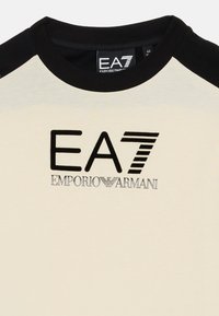 Krém színű póló fekete raglán ujjal. Az elején fekete és ezüst színű "EA7 EMPORIO ARMANI" logó található. Puha anyag, kerek nyakkivágással.