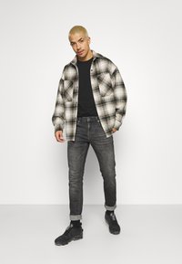 Overmaatse geruite shirt in crème, zwart en grijs, gedragen over een zwart t-shirt; gecombineerd met donkere jeans en zwarte sportschoenen.