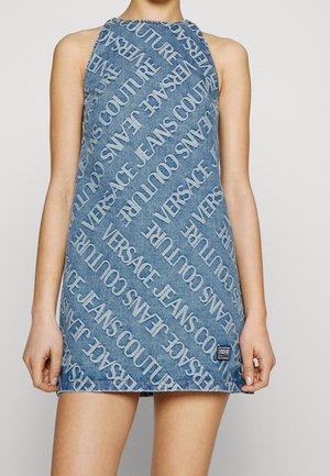 Femme portant une robe mini en denim bleu sans manches avec un motif répétitif en diagonale du texte "VERSACE JEANS COUTURE".