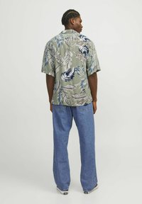 Camisa de manga corta verde con un patrón de hojas en azul y blanco, combinada con unos vaqueros claros sueltos. Fondo liso, vista desde la parte trasera.