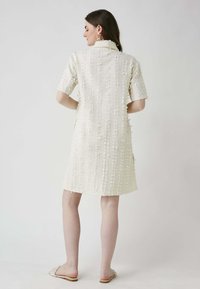 Weißes, strukturiertes Kleid mit kurzenÄrmeln, einem kragenförmigen Ausschnitt, gesticktem Muster und ungleichmäßigem Saum, kombiniert mit hellen, offenen Sandalen.