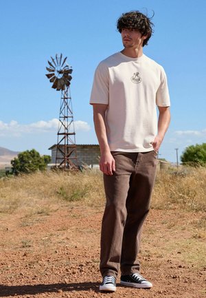 Muž oblečený v béžovém tričku Mack Trucks, hnědých kalhotách a černých teniskách stojí na suchém poli s větrným mlýnem a budovou v pozadí.