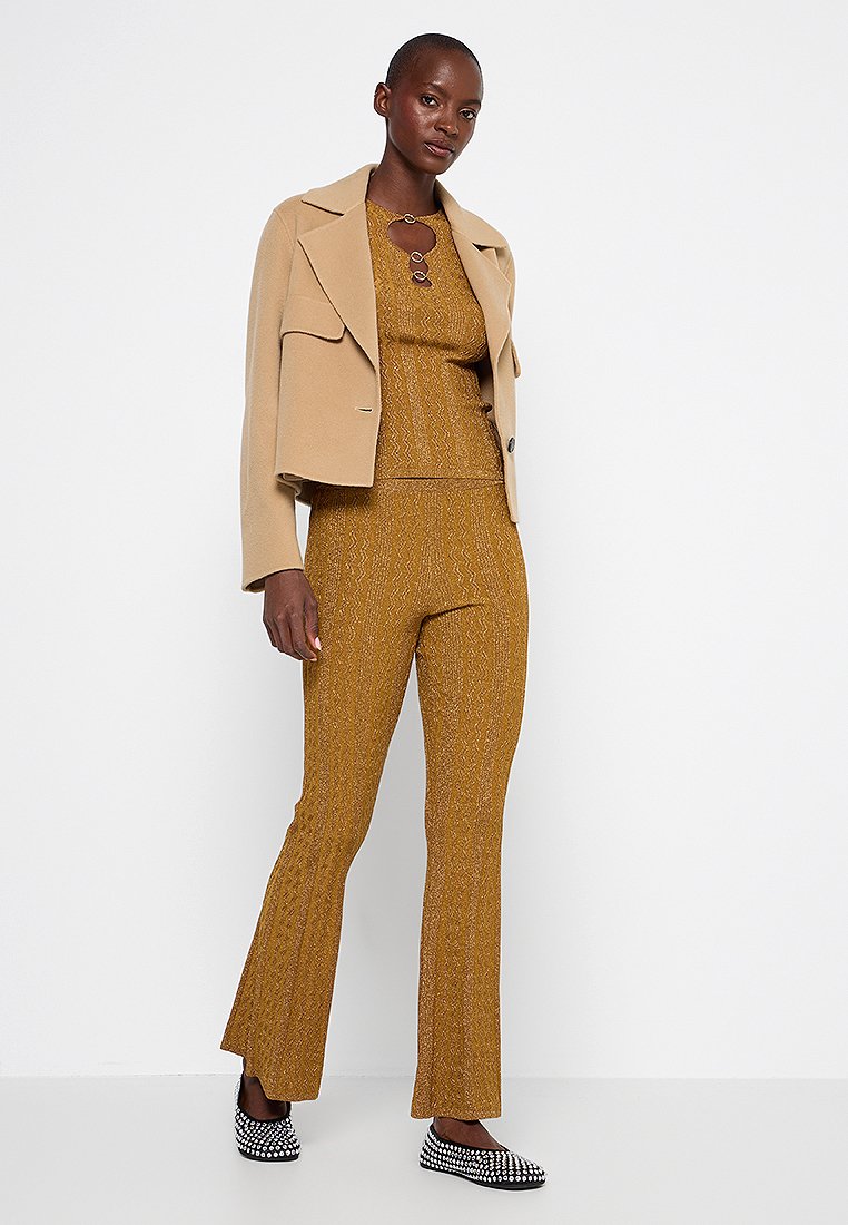 Beige kurzes Jackett über einem senfgelben Stricktop und ausgestellten Hosen, die ein strukturiertes Muster aufweisen. Schwarz-weiße karierte Schuhe runden den Look ab.