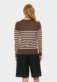 Donna bionda che indossa un maglione a righe marroni e crema con maniche a sbuffo e pantaloni corti neri fino al ginocchio, vista da dietro.