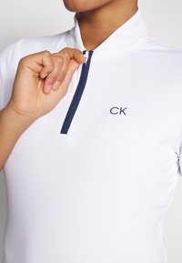 White polo shirt with a zip collar, featuring a navy zipper and CK logo on the chest. The fabric has a textured finish.