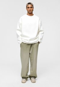 Witte oversized sweater met verlaagde schouders, gecombineerd met losse, licht olijfgroene broeken. Casual beige sneakers maken de outfit compleet.