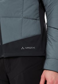 Giacca leggera in piumino grigio-blu, con motivo trapuntato e pannelli neri. Include una zip e il logo Vaude vicino all'orlo.