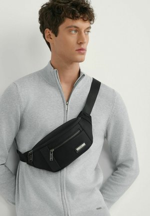 Młody mężczyzna w jasnoszarym swetrze zapinanym na zamek i czarnej torbie na pasku z dwoma komorami na zamek, spoglądający na bok.