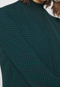 Donna che indossa un top a collo alto e maniche lunghe con un motivo geometrico zebbrato blu, verde e viola, con un orecchino a cerchio visibile.