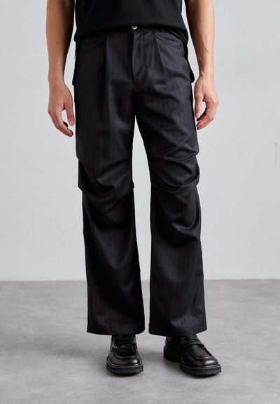 Pantalones de rayas negras con un corte holgado, que cuentan con bolsillos laterales y un diseño de pierna recta, combinados con zapatos negros y una camiseta negra.