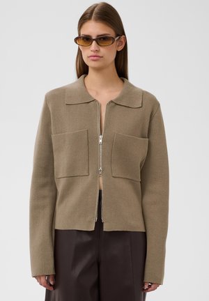 Femme portant un cardigan zippé marron avec de grandes poches devant, un pantalon marron foncé et des lunettes de soleil fines sur un fond clair uni.