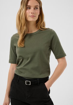 Femme aux longs cheveux blonds portant un t-shirt à manches courtes vert olive rentré dans un pantalon noir avec une ceinture noire, debout avec une main dans la poche.
