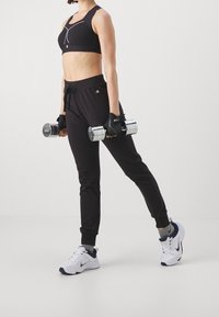 Black sports bra with white accents, black sweatpants with a drawstring, gray socks, white sneakers, and metallic dumbbells in hands.
