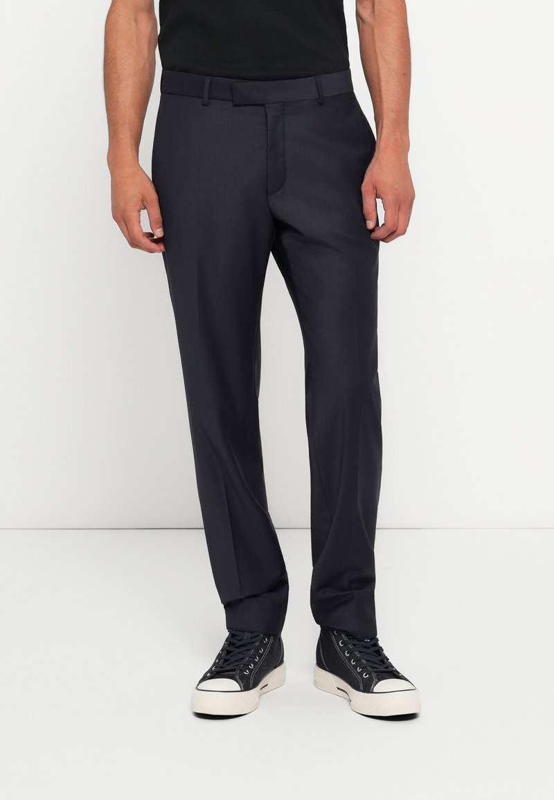 Calças sob medida cinza escuras com frente lisa, passadores de cinto e uma textura suave, combinadas com ténis pretos de cano alto e uma blusa preta.