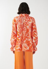 Blusa floreale in vivaci tonalità di arancione e rosa con maniche lunghe e polsini arricciati, abbinata a pantaloni arancioni a gamba larga.