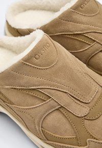 Mäkké kožené papuče v jemnej béžovej farbe s oblou šijacou líniou, širokým nastaviteľným pásikom a hrubou bielou flísovou podšívkou pre teplo a pohodlie.