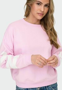 Helehele roosa dressipluus valgete kirjastega varrukatel, ümara kaelusega ja soonikutega mansettidega. Model kannab teksapükstega lühikesi pükse. Käed esiplaanil.