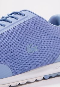 Světle modrá sportovní obuv s texturovaným síťovým svrškem, kontrastními hladkými akcenty a logem Lacoste. Obsahuje ploché tkaničky a bílou podrážku.