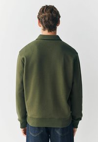 Grüner Pullover mit glatter Textur, der über einen Kragen, gerippte Bündchen und Saum sowie eine lässige Passform verfügt. Die Rückansicht zeigt die Nahtdetails.