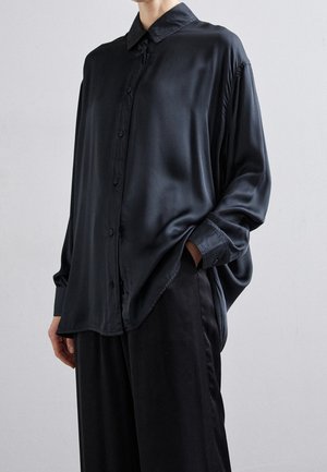 Personne portant une chemise ample en satin noir boutonnée et un pantalon noir assorti, une main dans la poche, sur un fond uni.