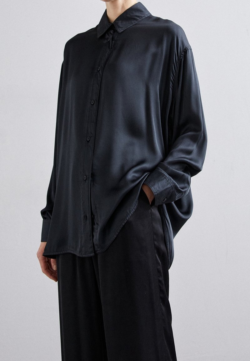 Personne portant une chemise ample en satin noir boutonnée et un pantalon noir assorti, une main dans la poche, sur un fond uni.