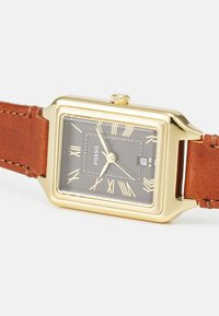 Montre rectangulaire dorée avec un cadran gris foncé, des chiffres romains et un bracelet en cuir marron avec des détails de couture.