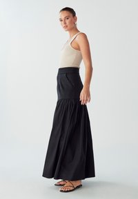 Gonna maxi nera con vita aderente, dettagli plissettati e orlo ampio; abbinata a una canotta beige a coste e sandali neri.